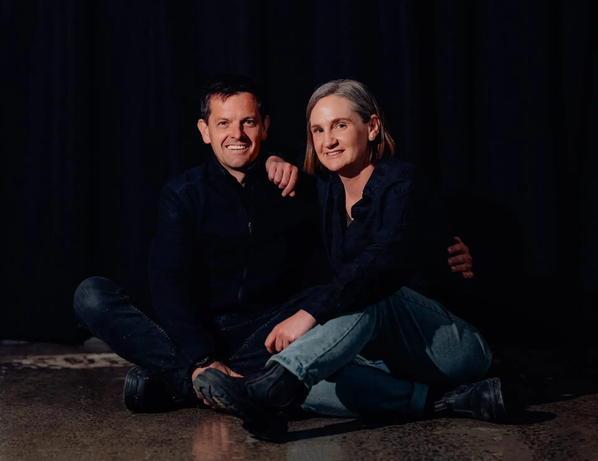 Co-owners and winemakers of Alchemy Wines, Cairn and Rebecca Coghill, together for a charity wine auction photo shoot, sitting on the floor in a wine cellar, smiling at the camera. Family owned